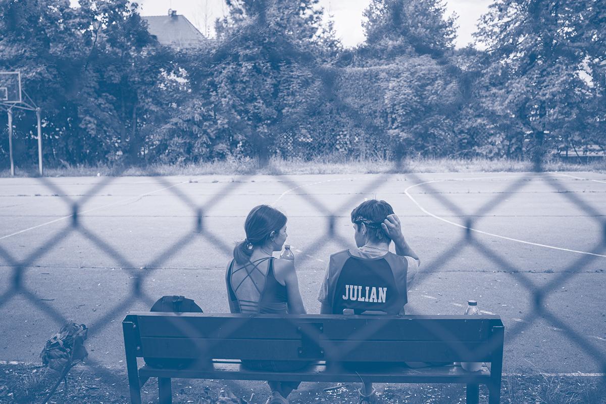 zdjęcie ukazujące dwoje dzieci siedzących na ławce, na boisku. widok zza siatki ogrodzeniowej. 