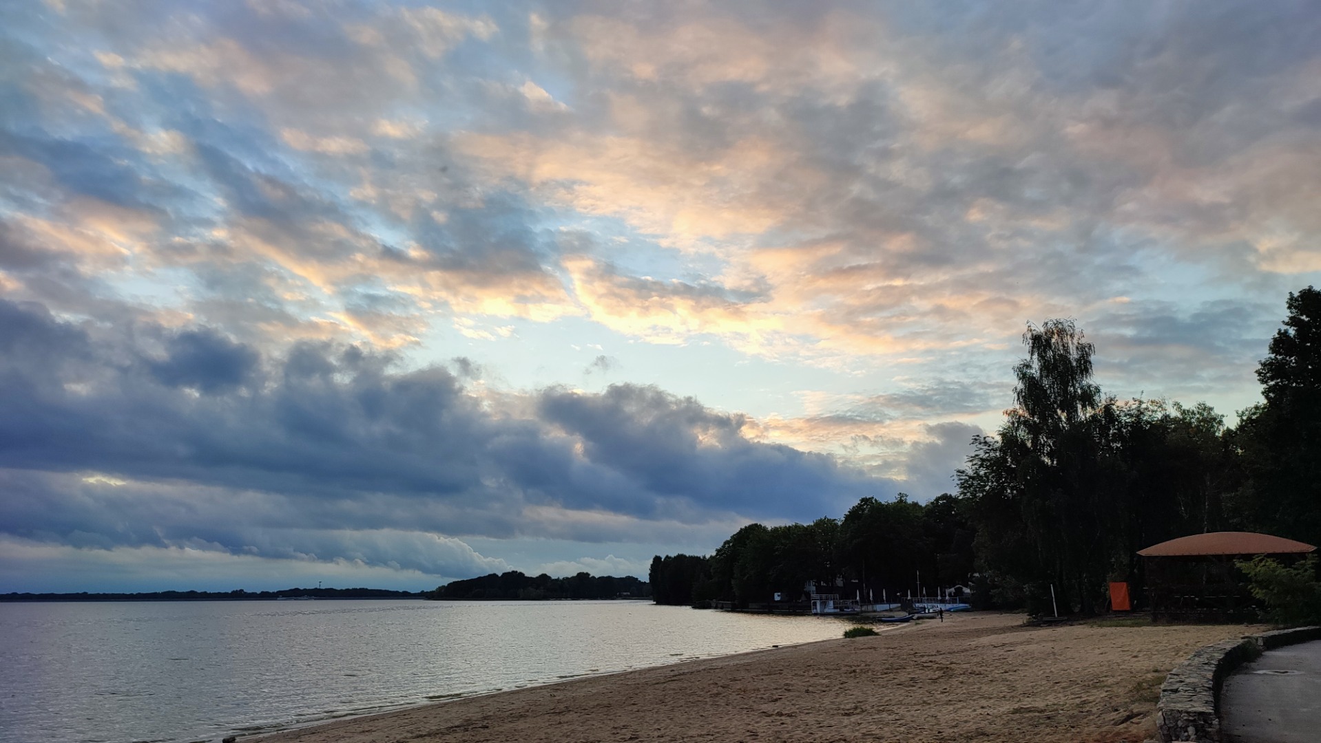 fotografia ukazująca zachód słońca nad jeziorem, fragment plaży z drzewami. 
